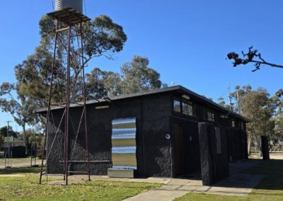 An exterior view of a rustic amenities block at Stawell Grampians Caravan Park, located near Stawell Hotel Motel Accommodation. The building has a dark, rugged facade with corrugated metal features, a modern design blending with the surrounding nature. A tall water tank is visible on a metal stand beside the structure, and trees and grassy areas surround the building under a clear blue sky