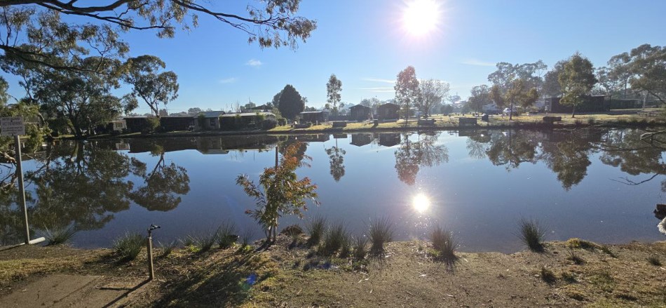 Serene view of a tranquil lake reflecting the bright sun and clear blue sky, framed by lush greenery and quaint cottages, showcasing the picturesque beauty of Stawell Hotel Motel Accommodation, an ideal destination for relaxation and nature exploration