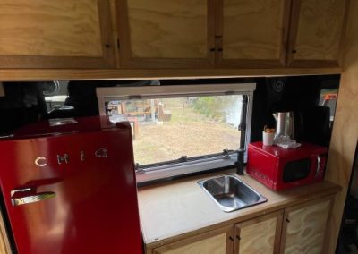 Compact kitchenette inside a Hammervan at Stawell Hotel Motel Accommodation, featuring wooden cabinetry, a red CHiQ mini-fridge, red microwave, sink, and kettle. A window above the sink provides a view of the outside, creating a cozy and functional space for guests