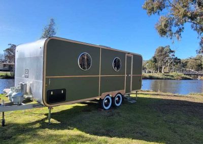 A green retro-style caravan with round windows, positioned on the grass beside the lake at Stawell Grampians Gate C&C Park – one of the options among Cabins, Dwellings, Caravans, & Glamping Tents.