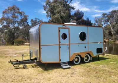Light blue retro caravan with timber trim parked beside a lake under a bright sky. One of the Cabins, Dwellings, Caravans, & Glamping Tents at Stawell Grampians Gate C&C Park.