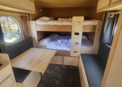 Interior of a light timber caravan with bunk beds, built-in table, and circular window. One of the Cabins, Dwellings, Caravans, & Glamping Tents at Stawell Grampians Gate C&C Park.
