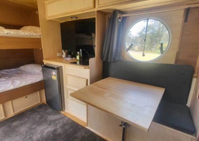 Compact dining nook inside a timber caravan with round window, bunk beds, and kitchenette. One of the Cabins, Dwellings, Caravans, & Glamping Tents at Stawell Grampians Gate C&C Park.