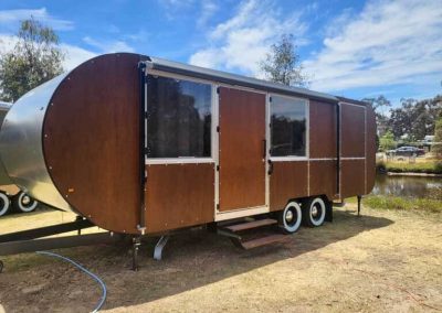 Sleek brown-panelled caravan with round ends and twin axles, set beside a pond. One of the Cabins, Dwellings, Caravans, & Glamping Tents at Stawell Grampians Gate C&C Park.