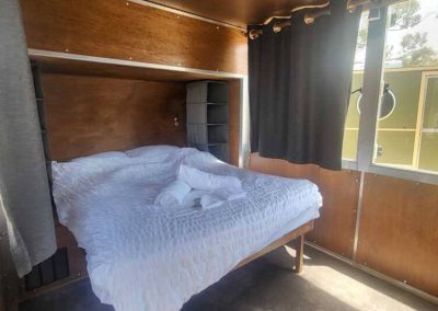 Double bed setup inside a brown caravan with storage shelves and natural light. One of the Cabins, Dwellings, Caravans, & Glamping Tents at Stawell Grampians Gate C&C Park.