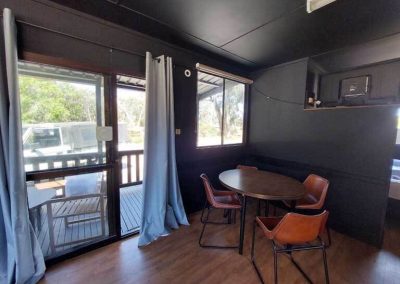 Dining nook with round wooden table and four leather chairs, adjacent to sliding doors leading to a covered deck. One of the Cabins, Dwellings, Caravans, & Glamping Tents at Stawell Grampians Gate C&C Park.