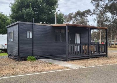 Dark-clad cabin with a small covered deck and timber railing, set within a natural campground. One of the Cabins, Dwellings, Caravans, & Glamping Tents at Stawell Grampians Gate C&C Park.