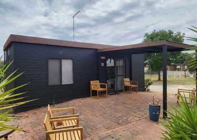 Front view of black cabin 53 with a large paved patio, covered entry, and wooden outdoor seating. One of the Cabins, Dwellings, Caravans, & Glamping Tents at Stawell Grampians Gate C&C Park.