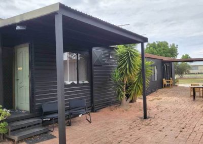 Black cabin with a sheltered porch, green front door, and paved outdoor seating area lined with tropical plants. One of the Cabins, Dwellings, Caravans, & Glamping Tents at Stawell Grampians Gate C&C Park