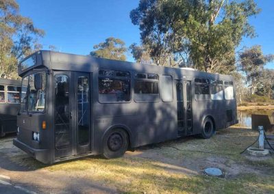 Converted black bus parked beside a lake under trees at Stawell Grampians Gate C&C Park. One of the Cabins, Dwellings, Caravans, & Glamping Tents at Stawell Grampians Gate C&C Park.