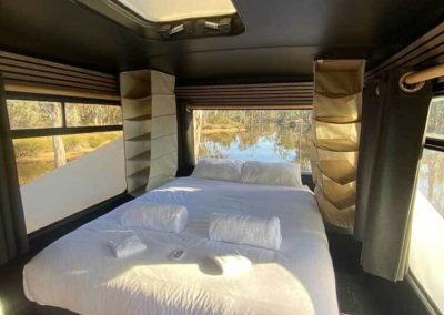 Double bed with fresh linens and a skylight above, surrounded by large windows overlooking the lake. One of the Cabins, Dwellings, Caravans, & Glamping Tents at Stawell Grampians Gate C&C Park.