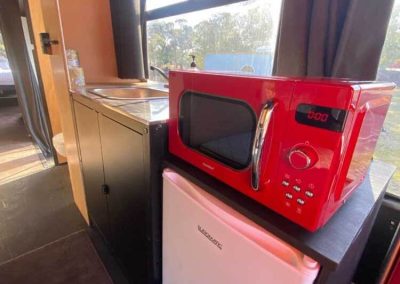 Compact kitchenette inside a converted bus with a red microwave, mini fridge, and stainless steel sink. One of the Cabins, Dwellings, Caravans, & Glamping Tents at Stawell Grampians Gate C&C Park.