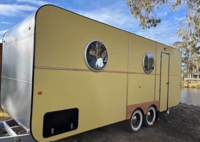 Yellow caravan with circular windows parked beside a lake at Stawell Grampians Gate C&C Park. One of the Cabins, Dwellings, Caravans, & Glamping Tents at Stawell Grampians Gate C&C Park.