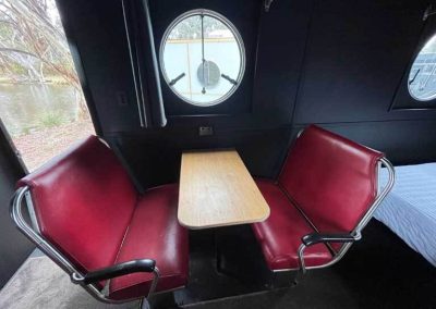 Interior of a retro caravan with round windows, a red retro-style fridge, compact dining area, and made-up double bed. One of the Cabins, Dwellings, Caravans, & Glamping Tents at Stawell Grampians Gate C&C Park.