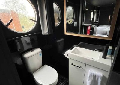 Compact bathroom with dark walls and round porthole-style windows. Includes a white toilet, white vanity with towel, and a large mirror reflecting the main room. One of the Cabins, Dwellings, Caravans, & Glamping Tents at Stawell Grampians Gate C&C Park.