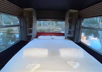 A double bed with white linen is set at the end of a converted bus, surrounded by large windows and shelving units on either side. The view outside shows water and trees. One of the Cabins, Dwellings, Caravans, & Glamping Tents at Stawell Grampians Gate C&C Park.