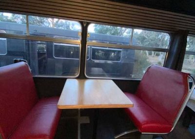 Converted bus with dark interior walls and metallic trim. Red vinyl booth seating with a wooden table set next to large bus windows. One of the Cabins, Dwellings, Caravans, & Glamping Tents at Stawell Grampians Gate C&C Park.