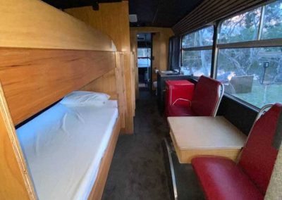 Interior view of a converted bus with wooden bunk beds on the left, a red microwave and small sink in the kitchenette on the right, and red diner-style booth seating. One of the Cabins, Dwellings, Caravans, & Glamping Tents at Stawell Grampians Gate C&C Park.