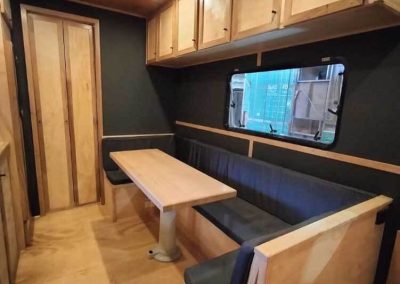 Interior of a retro van at Stawell Grampians Gate C&C Park showing a dining area with built-in bench seating, table, overhead cupboards, and window view – one of the Cabins, Dwellings, Caravans, & Glamping Tents available.