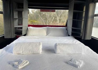 Interior view of a bed at the back of a converted bus, framed by windows on three sides with grey curtains and soft shelving. One of the Cabins, Dwellings, Caravans, & Glamping Tents at Stawell Grampians Gate C&C Park.