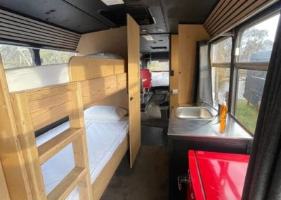 Interior of a converted bus showing a compact layout with wooden bunk beds on the left, a red mini fridge, and a small kitchenette with a sink on the right. Dining and sleeping areas are visible toward the rear. One of the Cabins, Dwellings, Caravans, & Glamping Tents at Stawell Grampians Gate C&C Park.
