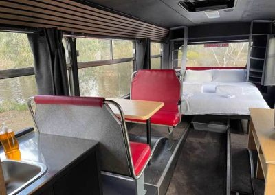 Interior of a converted bus featuring a booth-style dining setup with red seats, a kitchenette with sink and dish soap, and a raised sleeping area with a double bed at the rear. Large windows line the bus, offering views of the outside surroundings. One of the Cabins, Dwellings, Caravans, & Glamping Tents at Stawell Grampians Gate C&C Park.