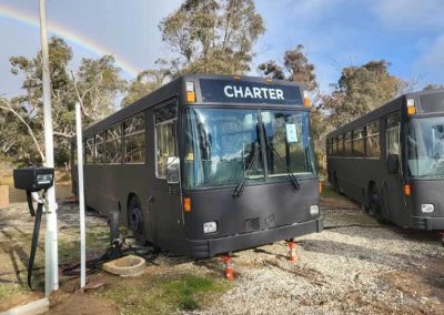 Two repurposed black charter buses parked on gravel, with a visible rainbow in the background and electrical connections running to the side. One of the Cabins, Dwellings, Caravans, & Glamping Tents at Stawell Grampians Gate C&C Park.