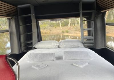 Double bed with white linen and towels positioned at the rear of a bus conversion, surrounded by windows and built-in shelving. View outside includes water and trees. One of the Cabins, Dwellings, Caravans, & Glamping Tents at Stawell Grampians Gate C&C Park.