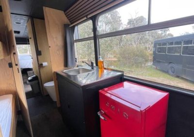 Kitchenette inside a converted bus featuring a stainless steel sink with soap dispensers, a compact red refrigerator, and large windows providing natural light. A door leads to the bathroom area. One of the Cabins, Dwellings, Caravans, & Glamping Tents at Stawell Grampians Gate C&C Park.