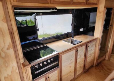 Interior view of a retro van kitchen, featuring a compact gas stove with oven, small sink, and light wood cabinetry, available at Stawell Grampians Gate C&C Park – one of the Cabins, Dwellings, Caravans, & Glamping Tents.