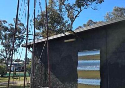 A tall water tank on a metal frame stands beside a dark-colored building with a shiny corrugated metal feature on its wall, under a clear blue sky. One of the features at Stawell Grampians Gate C&C Park – Cabins, Dwellings, Caravans, & Glamping Tents.