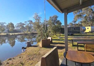 Covered seating area beside the lake with wooden tables and benches. Retro caravans and trees in the background. One of the Cabins, Dwellings, Caravans, & Glamping Tents at Stawell Grampians Gate C&C Park.
