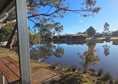 Bench seating under a shelter facing the lake. Still water reflects trees and buildings across the shore. One of the Cabins, Dwellings, Caravans, & Glamping Tents at Stawell Grampians Gate C&C Park.