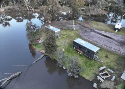 Aerial view of two retro vans parked along the water’s edge at Stawell Grampians Gate C&C Park, each with nearby fire pits and surrounded by natural bushland – one of the Cabins, Dwellings, Caravans, & Glamping Tents available.