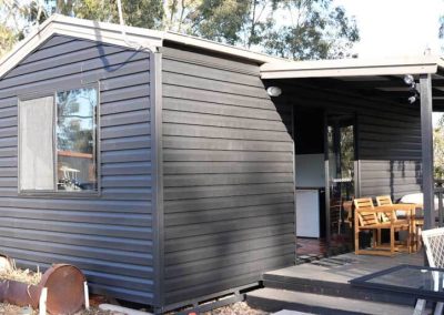 Exterior view of a black weatherboard cabin with a covered deck and outdoor seating, located beside the lake at Stawell Grampians Gate C&C Park. One of the Cabins, Dwellings, Caravans, & Glamping Tents available at the park.