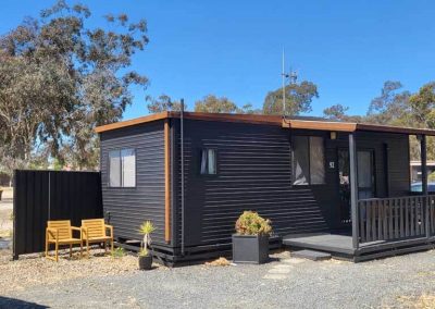 A black rectangular cabin with vertical corrugated metal siding, a flat roof, and timber trim. A small covered porch with a railing and steps leads to the front door marked “92.” There are two outdoor wooden chairs and a table placed on a gravel area to the side, and potted plants add greenery around the entrance. One of the Cabins, Dwellings, Caravans, & Glamping Tents at Stawell Grampians Gate C&C Park.