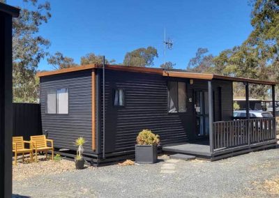 A black cabin with a flat roof and timber trim, featuring a small front porch with railing and a gravel path leading to the entrance. Two wooden chairs sit beside the building on a patch of decorative gravel, and a potted plant is placed near the steps. Trees and other cabins are visible in the background. One of the Cabins, Dwellings, Caravans, & Glamping Tents at Stawell Grampians Gate C&C Park.