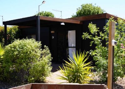 A compact black cabin marked with the number 54, surrounded by dense green plants and shrubs, with a small covered entryway. One of the Cabins, Dwellings, Caravans, & Glamping Tents at Stawell Grampians Gate C&C Park.