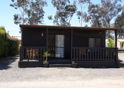 A small black cabin with a covered front deck, outdoor seating, potted plants, and gravel surroundings. One of the Cabins, Dwellings, Caravans, & Glamping Tents at Stawell Grampians Gate C&C Park.