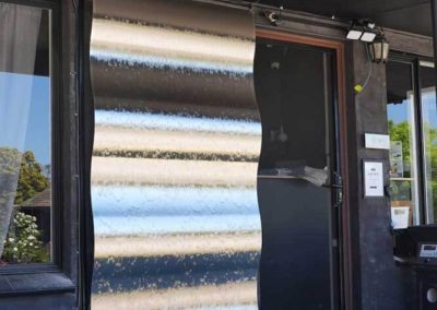 A vertical corrugated metal water feature is mounted beside a dark-framed glass door. Water trickles down the reflective surface into a rectangular basin filled with dark stones. One of the Cabins, Dwellings, Caravans, & Glamping Tents at Stawell Grampians Gate C&C Park.