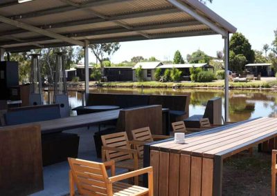 Covered outdoor seating area with wooden tables and a mix of timber and padded bench seating, set beside a waterway. One of the Cabins, Dwellings, Caravans, & Glamping Tents at Stawell Grampians Gate C&C Park.