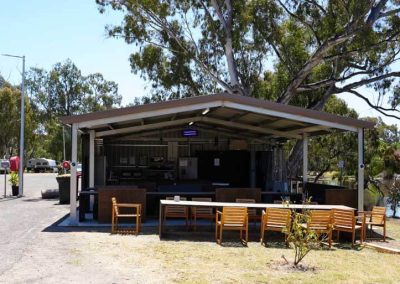 A covered outdoor kitchen and dining area with wooden tables and chairs arranged in front. Inside, there are metal countertops, a fridge, and cooking appliances under a corrugated metal roof supported by poles. The space is surrounded by trees, with a pond visible to the right. One of the Cabins, Dwellings, Caravans, & Glamping Tents at Stawell Grampians Gate C&C Park.