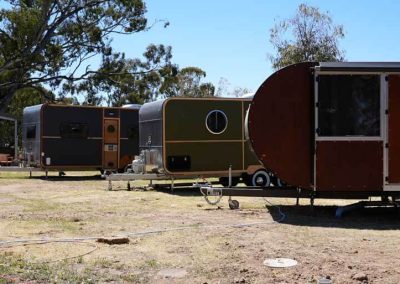Three caravans of different shapes and colours arranged in a line. The brown caravan with rounded ends is in the foreground, followed by a green one with a circular window. Set on dry grass under trees. One of the Cabins, Dwellings, Caravans, & Glamping Tents at Stawell Grampians Gate C&C Park.