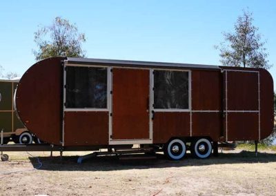 A brown caravan with a distinctive rounded shape on both ends, metal framing, and white-rimmed wheels. Positioned on a grassy area with a pond nearby. One of the Cabins, Dwellings, Caravans, & Glamping Tents at Stawell Grampians Gate C&C Park.