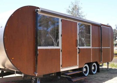 A brown caravan with rounded edges and multiple rectangular panels along its side, including two windows and two central doors. The van is set up on dry grass with a few trees and water visible in the background. One of the Cabins, Dwellings, Caravans, & Glamping Tents at Stawell Grampians Gate C&C Park.
