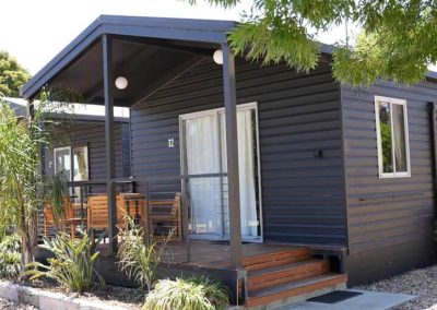 A dark-painted cabin with a small covered porch, outdoor seating, and wood steps leading up to a glass sliding door. Marked with the number 32 and surrounded by light landscaping. One of the Cabins, Dwellings, Caravans, & Glamping Tents at Stawell Grampians Gate C&C Park.