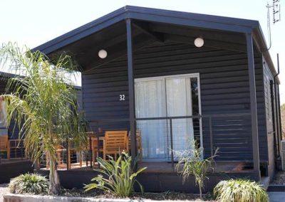 A compact black cabin with a gabled roof and a small front deck featuring a wooden outdoor dining set. Number 32 is displayed next to a sliding glass door, and low plants grow around the base. One of the Cabins, Dwellings, Caravans, & Glamping Tents at Stawell Grampians Gate C&C Park.