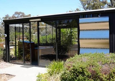 A glass-walled structure with sliding doors stands surrounded by low shrubs and native plants. Inside are long wooden tables and built-in bench seating. One of the Cabins, Dwellings, Caravans, & Glamping Tents at Stawell Grampians Gate C&C Park.