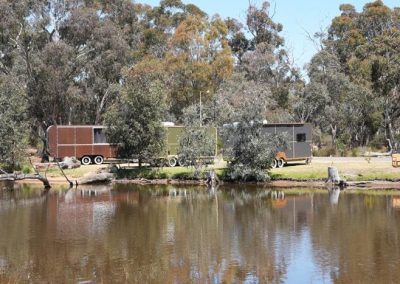 Three caravans with rounded ends are parked side-by-side behind a line of trees on the opposite bank of a calm lake. The reflection of the trees and caravans is visible in the water. One of the Cabins, Dwellings, Caravans, & Glamping Tents at Stawell Grampians Gate C&C Park.