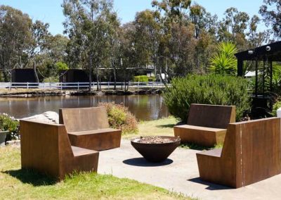 Four wooden benches are arranged around a round fire pit near the edge of a lake. A white fence and trees are visible across the water. One of the Cabins, Dwellings, Caravans, & Glamping Tents at Stawell Grampians Gate C&C Park.